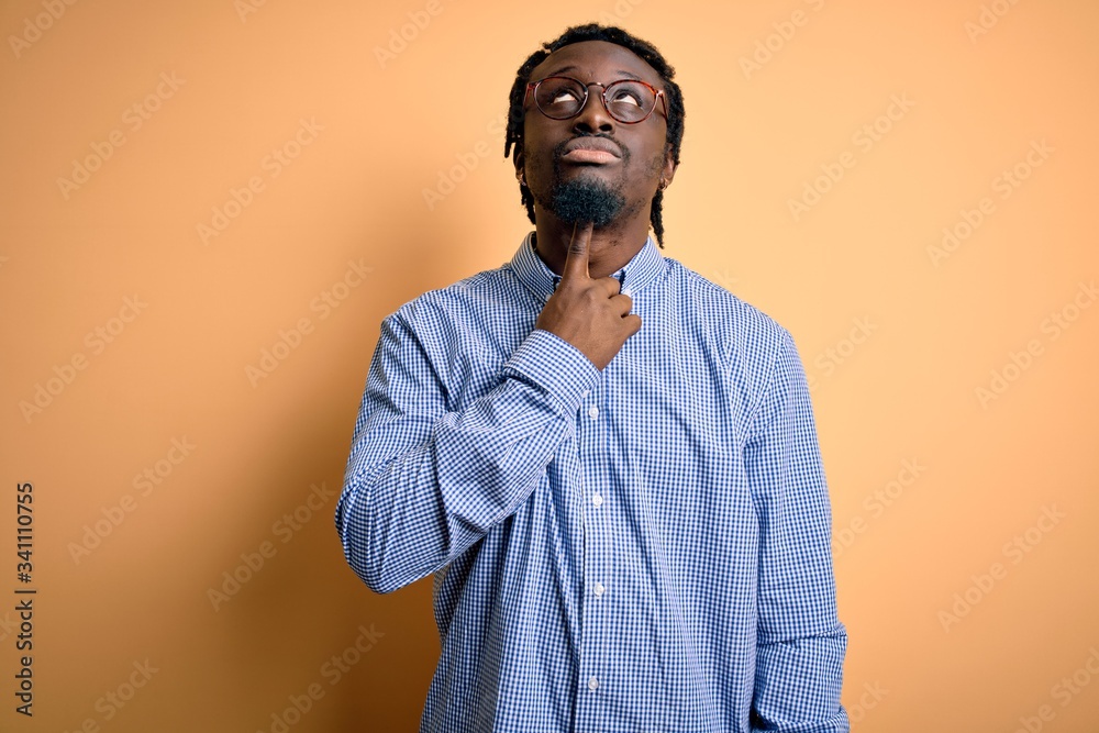Young handsome african american man wearing shirt and glasses over yellow background Thinking concentrated about doubt with finger on chin and looking up wondering Young handsome african american man wearing shirt and glasses over yellow background Thinking concentrated about doubt with finger on chin and looking up wondering
