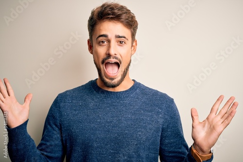 Young handsome man with beard wearing casual sweater standing over white background celebrating crazy and amazed for success with arms raised and open eyes screaming excited. Winner concept