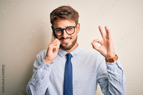 Young businessman having a conversation talking by smartphone over white background doing ok sign with fingers, excellent symbol