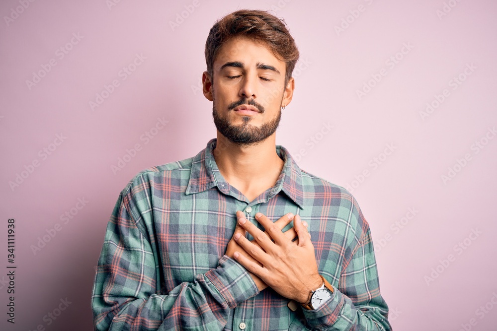Young handsome man with beard wearing casual shirt standing over pink background smiling with hands on chest with closed eyes and grateful gesture on face. Health concept. Young handsome man with beard wearing casual shirt standing over pink background smiling with hands on chest with closed eyes and grateful gesture on face. Health concept.