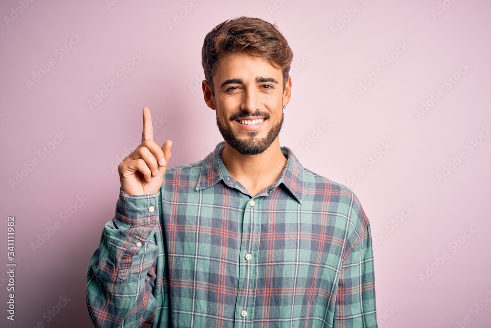 Young handsome man with beard wearing casual shirt standing over pink background showing and pointing up with finger number one while smiling confident and happy.