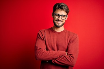 Young handsome man with beard wearing glasses and sweater standing over red background happy face smiling with crossed arms looking at the camera. Positive person.