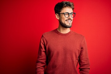 Young handsome man with beard wearing glasses and sweater standing over red background looking away to side with smile on face, natural expression. Laughing confident.