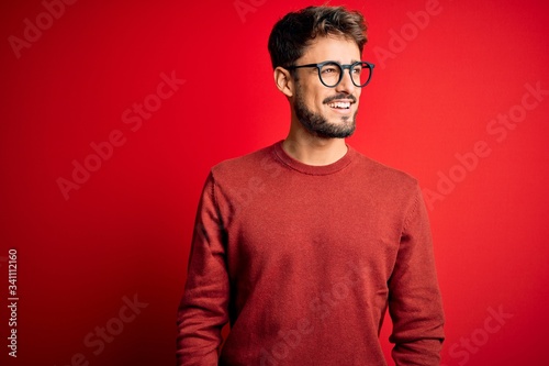 Young handsome man with beard wearing glasses and sweater standing over red background looking away to side with smile on face, natural expression. Laughing confident.