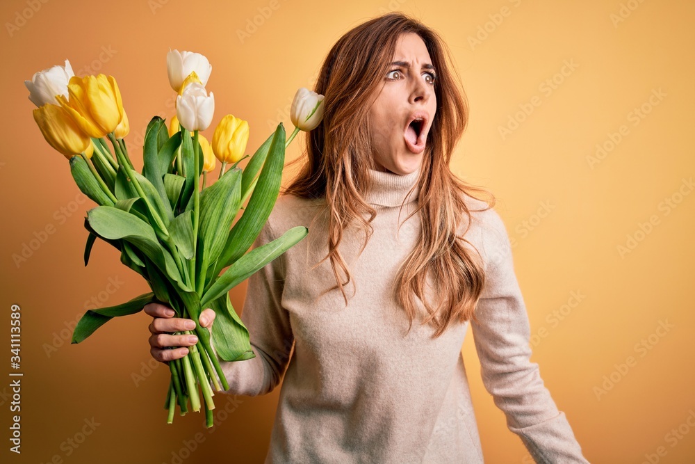 Young beautiful brunette woman holding bouquet of yellow tulips over isolated background angry and mad screaming frustrated and furious, shouting with anger. Rage and aggressive concept.