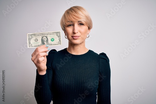 Young blonde woman with short hair holding one dollar banknote over isolated background with a confident expression on smart face thinking serious