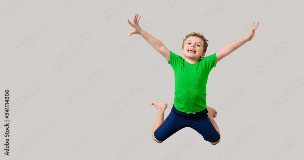 Happy little kid boy in green t-shirt on gray background is smiling, laughing and jumping
