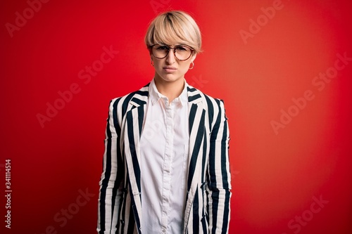 Blonde business woman with short hair wearing glasses and striped jacket over red background skeptic and nervous, frowning upset because of problem. Negative person.