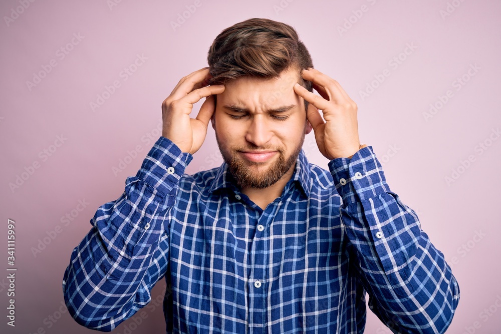Young handsome blond man with beard and blue eyes wearing casual shirt standing suffering from headache desperate and stressed because pain and migraine. Hands on head. Young handsome blond man with beard and blue eyes wearing casual shirt standing suffering from headache desperate and stressed because pain and migraine. Hands on head.