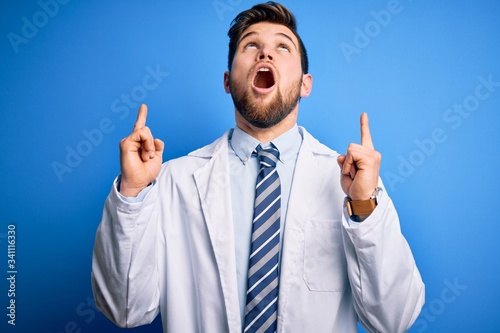 Young blond therapist man with beard and blue eyes wearing coat and tie over background amazed and surprised looking up and pointing with fingers and raised arms.
