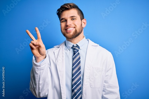 Young blond therapist man with beard and blue eyes wearing coat and tie over background showing and pointing up with fingers number two while smiling confident and happy.