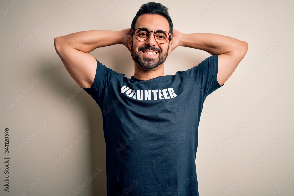 Handsome man with beard wearing t-shirt with volunteer message over white background relaxing and stretching, arms and hands behind head and neck smiling happy Handsome man with beard wearing t-shirt with volunteer message over white background relaxing and stretching, arms and hands behind head and neck smiling happy