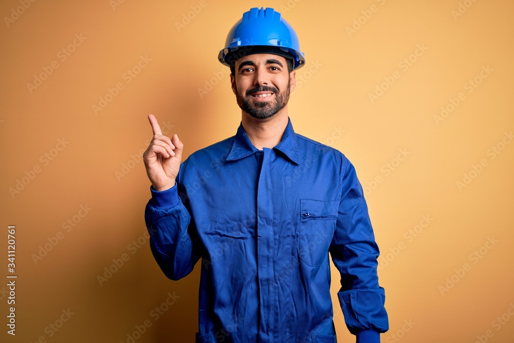Mechanic man with beard wearing blue uniform and safety helmet over yellow background with a big smile on face, pointing with hand finger to the side looking at the camera.