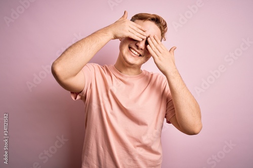 Young handsome redhead man wearing casual t-shirt standing over isolated pink background covering eyes with hands smiling cheerful and funny. Blind concept.
