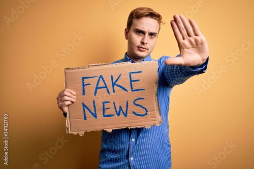 Canvas Print Young handsome redhead man holding banner with fake news message over yellow bac
