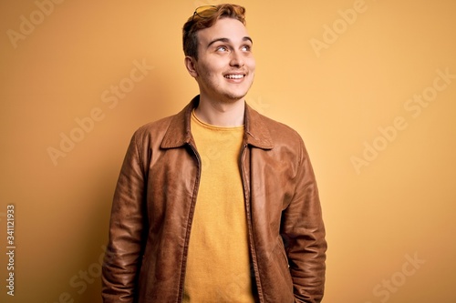 Young handsome redhead man wearing casual leather jacket over isolated yellow background smiling looking to the side and staring away thinking.