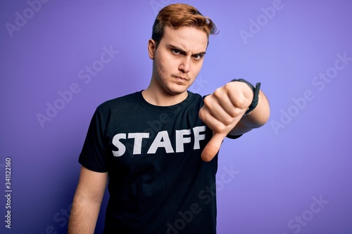 Young handsome redhead worker man wearing staff t-shirt uniform over purple background looking unhappy and angry showing rejection and negative with thumbs down gesture. Bad expression.