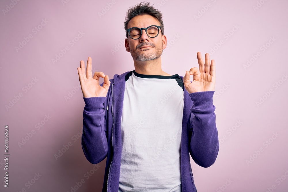 Young handsome man wearing purple sweatshirt and glasses standing over pink background relaxed and smiling with eyes closed doing meditation gesture with fingers. Yoga concept. Young handsome man wearing purple sweatshirt and glasses standing over pink background relaxed and smiling with eyes closed doing meditation gesture with fingers. Yoga concept.