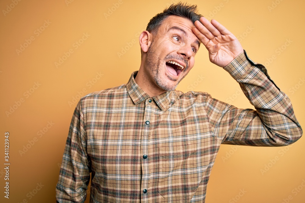 Young handsome man wearing casual shirt standing over isolated yellow background very happy and smiling looking far away with hand over head. Searching concept.