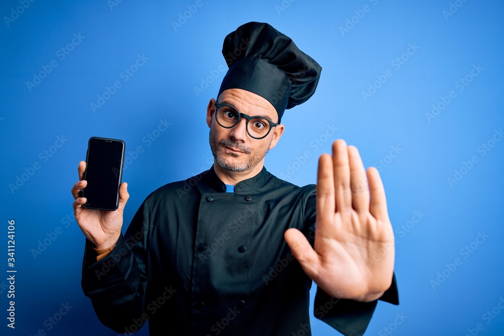 Young handsome chef man wearing uniform and hat holding smartphone over blue background with open hand doing stop sign with serious and confident expression, defense gesture Young handsome chef man wearing uniform and hat holding smartphone over blue background with open hand doing stop sign with serious and confident expression, defense gesture