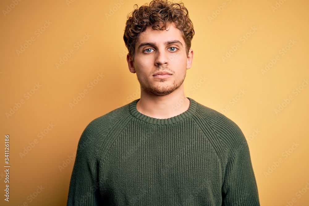 Young blond handsome man with curly hair wearing green sweater over yellow background with serious expression on face. Simple and natural looking at the camera.
