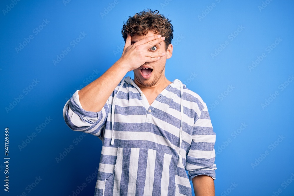 Young blond handsome man with curly hair wearing casual striped sweatshirt peeking in shock covering face and eyes with hand, looking through fingers with embarrassed expression. Young blond handsome man with curly hair wearing casual striped sweatshirt peeking in shock covering face and eyes with hand, looking through fingers with embarrassed expression.