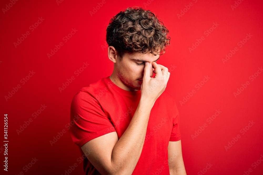 Young blond handsome man with curly hair wearing casual t-shirt over red background tired rubbing nose and eyes feeling fatigue and headache. Stress and frustration concept. Young blond handsome man with curly hair wearing casual t-shirt over red background tired rubbing nose and eyes feeling fatigue and headache. Stress and frustration concept.