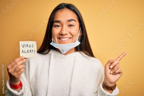 Young beautiful asian woman wearing medical mask holding paper with virus alert message very happy pointing with hand and finger to the side