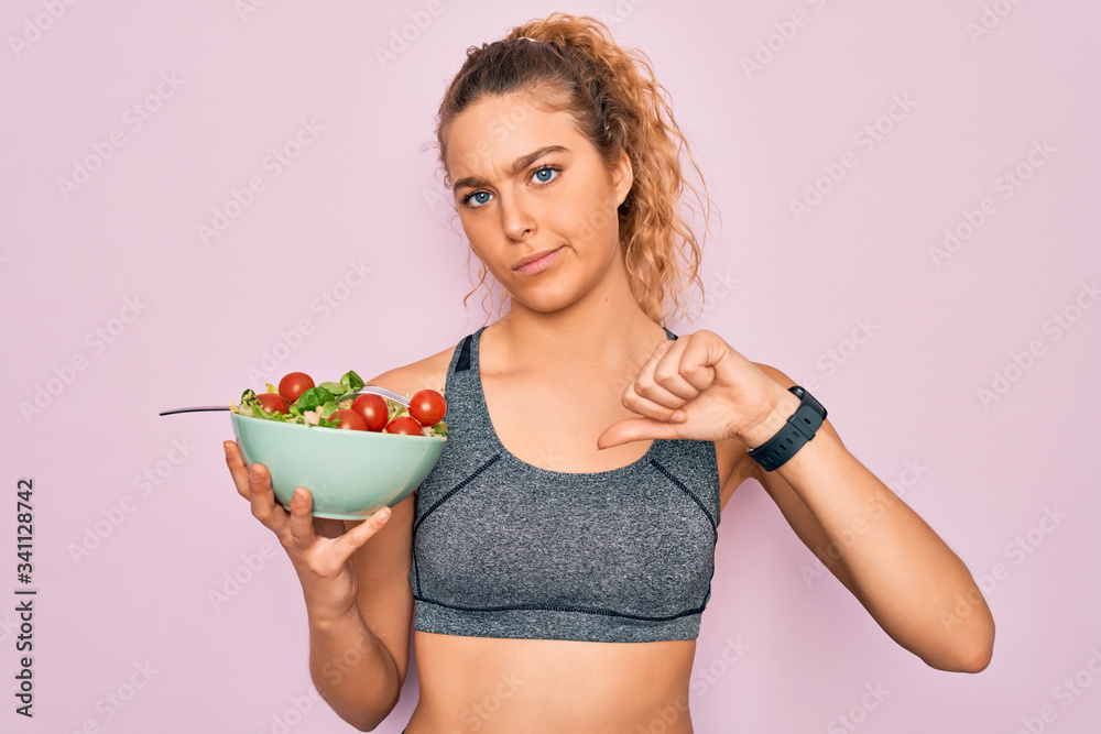 Young beautiful blonde sporty woman with blue eyes holding bowl with healthy salad with angry face, negative sign showing dislike with thumbs down, rejection concept