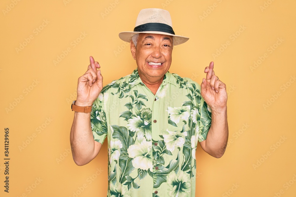 Middle age senior grey-haired man wearing summer hat and floral shirt on beach vacation gesturing finger crossed smiling with hope and eyes closed. Luck and superstitious concept.