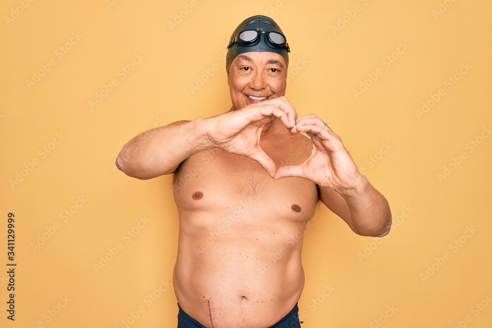 Middle age senior grey-haired swimmer man wearing swimsuit, cap and goggles smiling in love doing heart symbol shape with hands. Romantic concept.