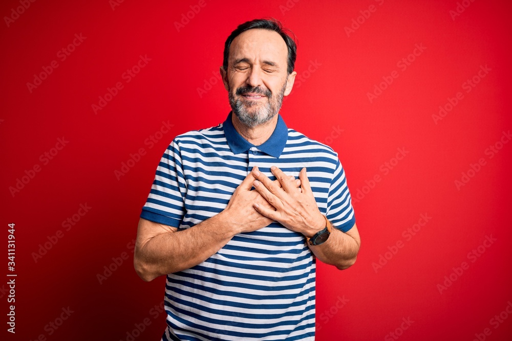 Middle age hoary man wearing casual striped polo standing over isolated red background smiling with hands on chest with closed eyes and grateful gesture on face. Health concept. Middle age hoary man wearing casual striped polo standing over isolated red background smiling with hands on chest with closed eyes and grateful gesture on face. Health concept.