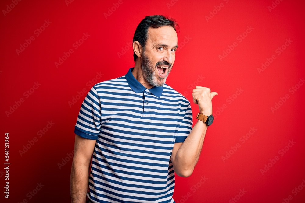 Fototapeta premium Middle age hoary man wearing casual striped polo standing over isolated red background smiling with happy face looking and pointing to the side with thumb up.