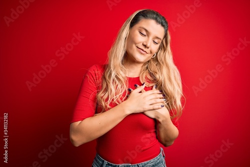 Young beautiful blonde woman wearing casual t-shirt standing over isolated red background smiling with hands on chest with closed eyes and grateful gesture on face. Health concept.