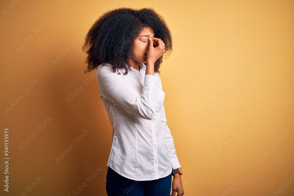 Young beautiful african american elegant woman with afro hair standing over yellow background tired rubbing nose and eyes feeling fatigue and headache. Stress and frustration concept. Young beautiful african american elegant woman with afro hair standing over yellow background tired rubbing nose and eyes feeling fatigue and headache. Stress and frustration concept.