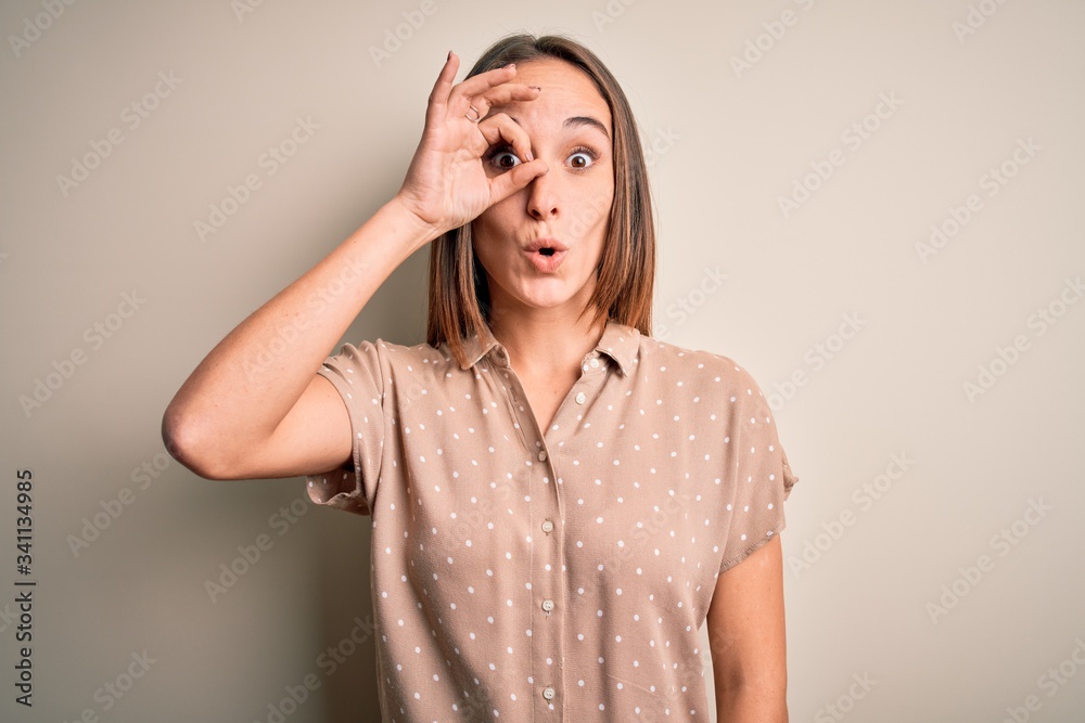 Young beautiful woman wearing casual shirt standing over isolated white background doing ok gesture shocked with surprised face, eye looking through fingers. Unbelieving expression. Young beautiful woman wearing casual shirt standing over isolated white background doing ok gesture shocked with surprised face, eye looking through fingers. Unbelieving expression.