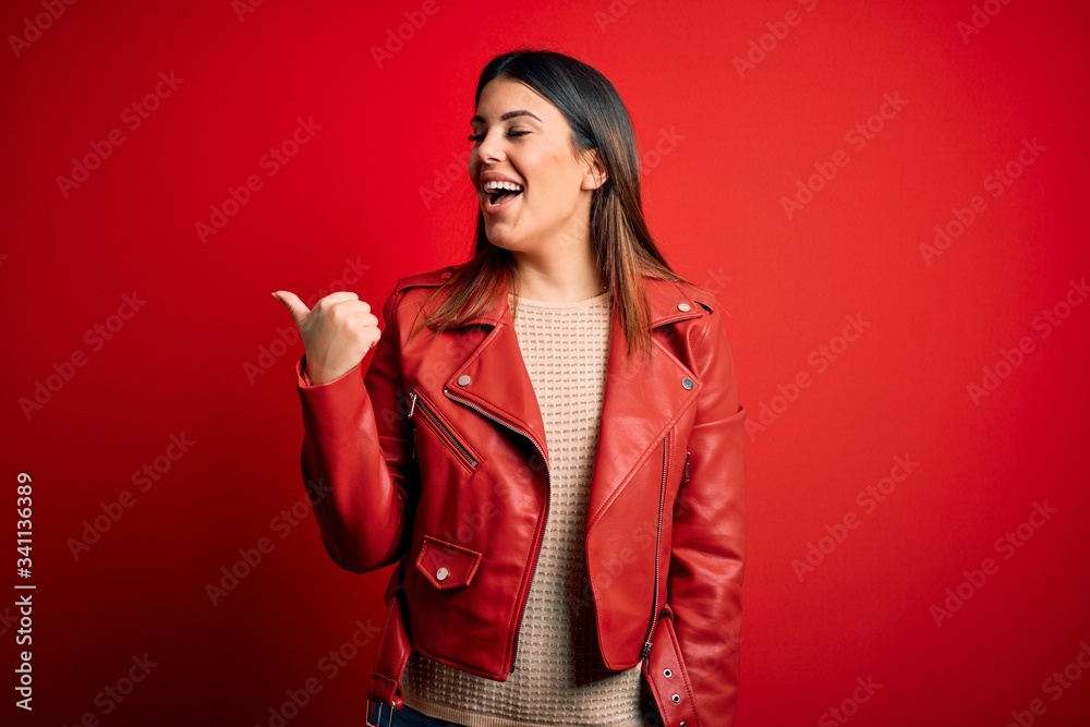 Young beautiful woman wearing red leather jacket over isolated background smiling with happy face looking and pointing to the side with thumb up.
