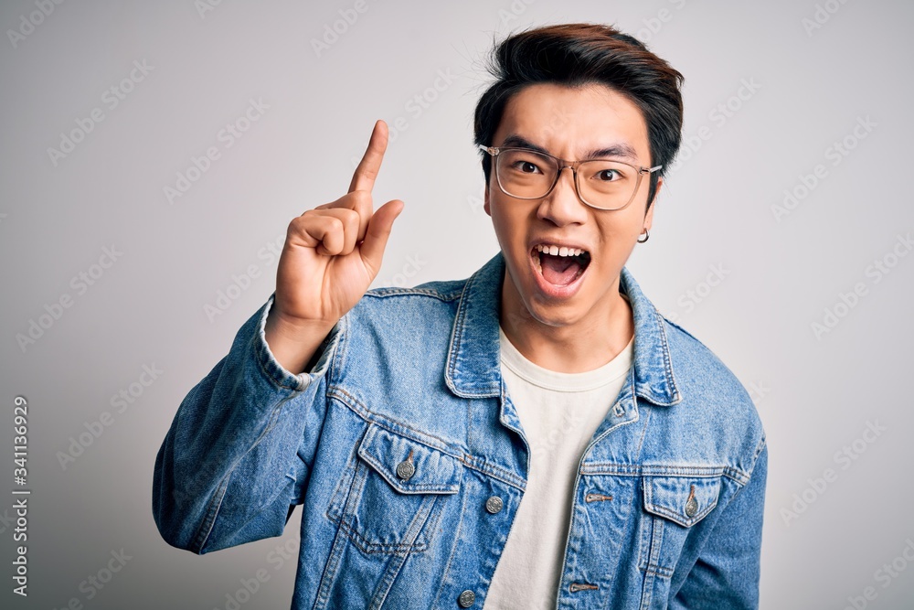 Young handsome chinese man wearing denim jacket and glasses over white background pointing finger up with successful idea. Exited and happy. Number one.