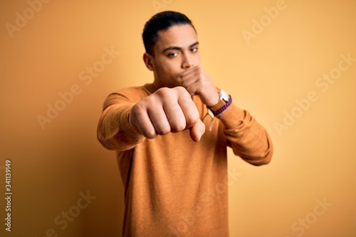 Young brazilian man wearing casual sweater standing over isolated yellow background Punching fist to fight, aggressive and angry attack, threat and violence