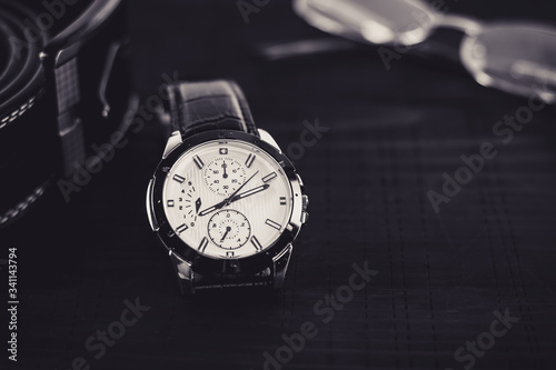 Elegant wristwatch on the wooden table with leather belt and glasses in the background. Close up, front view, male accessories concept.