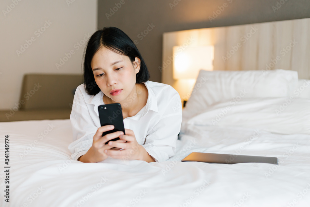 Beautiful asian female in white shirt using mobile phone while working on bed. work from home concept.