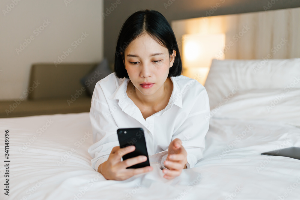 Beautiful asian female in white shirt using mobile phone while working on bed. work from home concept. Beautiful asian female in white shirt using mobile phone while working on bed. work from home concept.