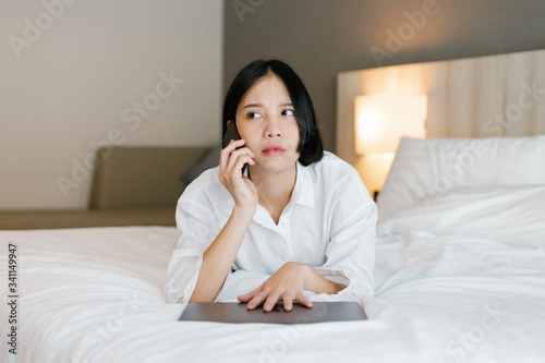 Beautiful asian female in white shirt using mobile phone while working on bed. work from home concept.