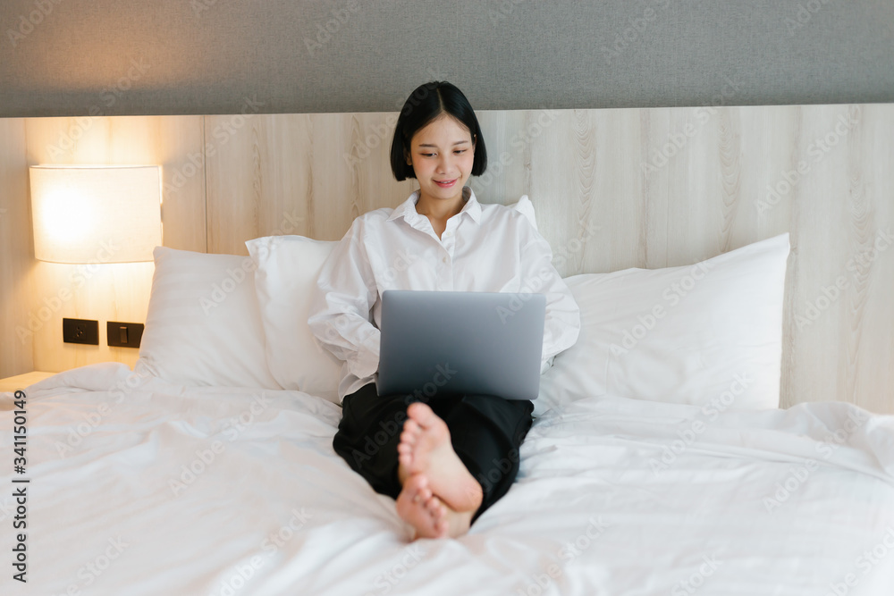 Beautiful asian female in white shirt using laptop while working on bed. work from home concept.
