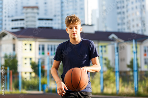 Cute smiling boy in blue t shirt plays basketball on city playground. Active teen enjoying outdoor game with orange ball. Hobby, active lifestyle, sport for kids.