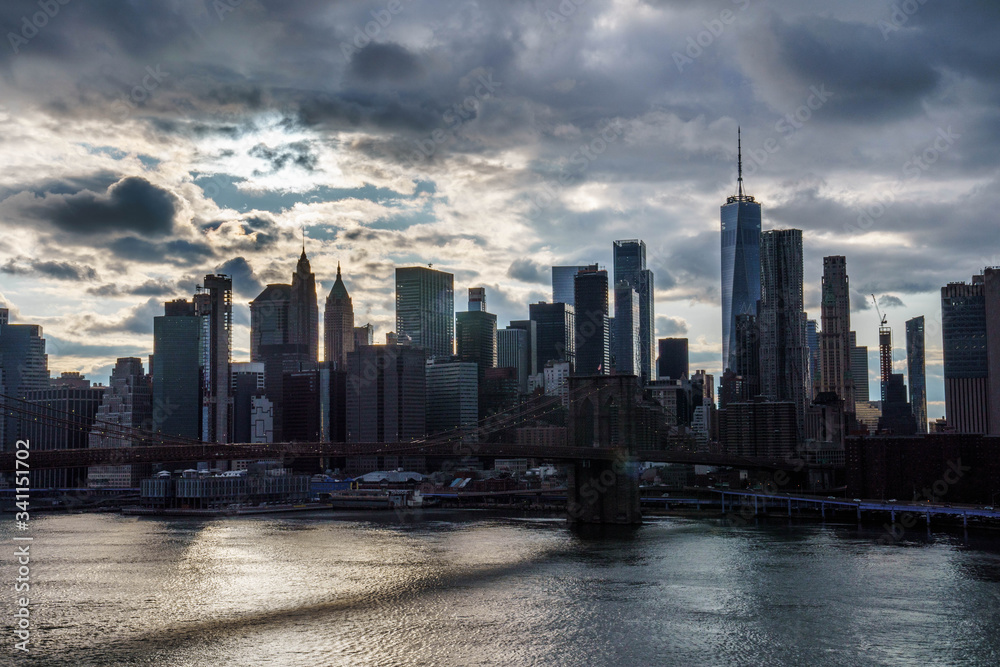 Manhattan skyline with Brooklyn bridge. Sky-rise skyscrapers tall apartment buildings. Dramatic cloudy sky with sun rays. 