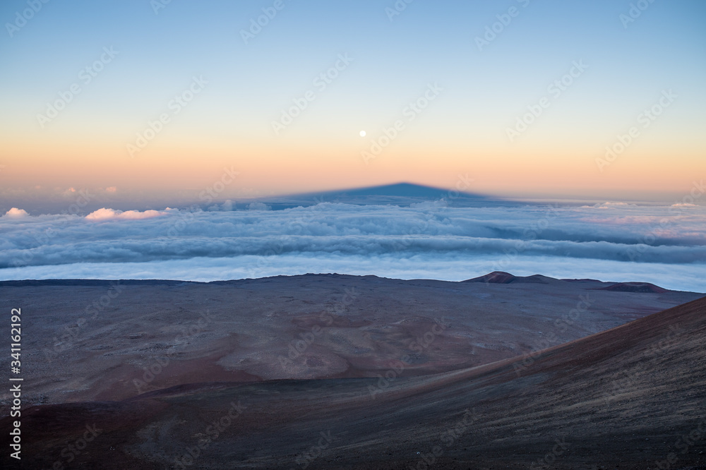 Shadow of the Mauna Kea volcano above clouds and near the moon.