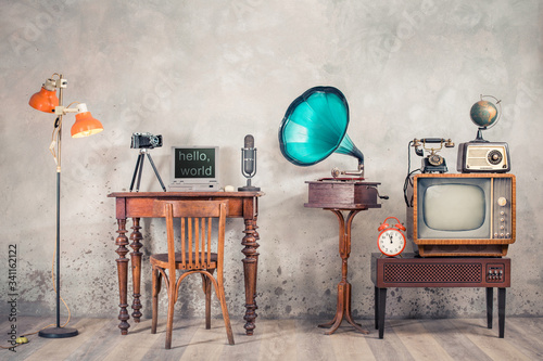 Retro TV, old radio, telephone, alarm clock, globe, gramophone, classic microphone, outdated laptop and camera on wooden table, floor lamp front concrete wall background. Vintage style filtered photo photo