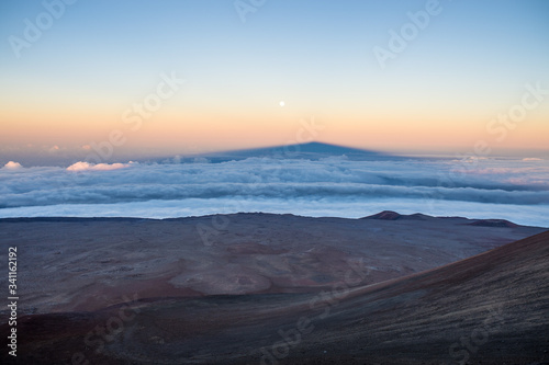 Shadow of the Mauna Kea volcano above clouds and near the moon.