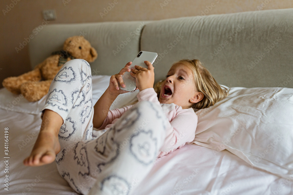 Cute little kid girl watching video on smartphone with smiley face alone on the bed, child using mobile phone with happy face at home. Stay at home quarantine coronavirus COVID-19 pandemic prevention. Cute little kid girl watching video on smartphone with smiley face alone on the bed, child using mobile phone with happy face at home. Stay at home quarantine coronavirus COVID-19 pandemic prevention.